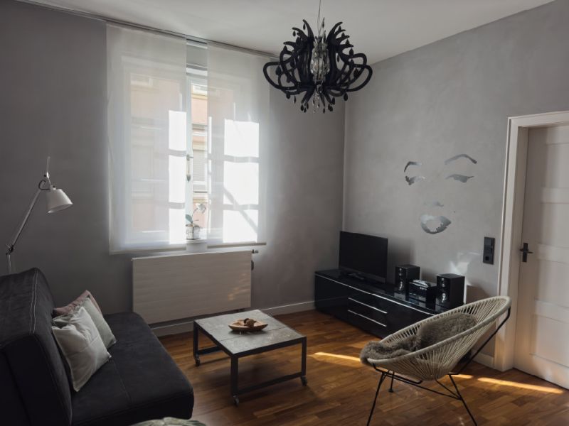 Helles Wohnzimmer der Ferienwohnung mit hellgrauen Wänden, schwarzem Kronleuchter, gemütlicher Sitzgelegenheit, Fernseher und hochwertigem Parkettboden im Altbau-Ambiente.