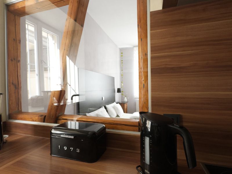 Moderne Küche der Ferienwohnung mit Holzarbeitsplatte, Wasserkocher und Blick durch ein Innenfenster in das gemütliche Schlafzimmer mit sichtbaren Holzbalken.